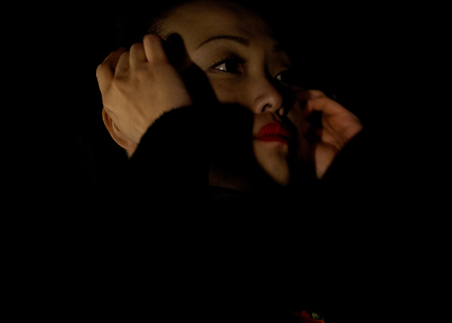 IMG_3371-2 A very close up image of a woman's face, mostly cast in shadow. Her hands are tucking hair being her ears and she wears red lipstick and looks off to teh right.